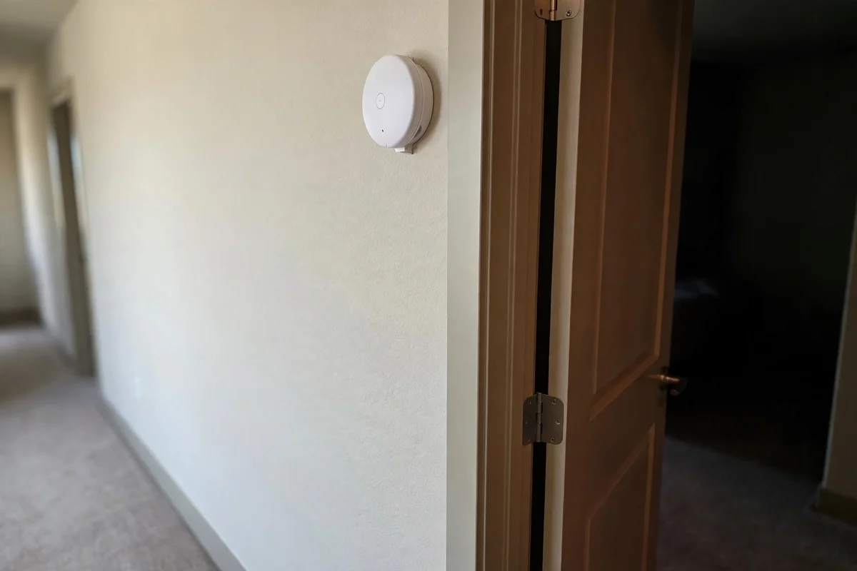 A plain white alarm mounted on a hallway wall near a bedroom door